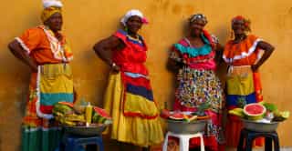 Vier trotse vrouwen in kleurrijke traditionele kledij verkopen vers fruit tegen een warmgele muur, een glimp van de levendige cultuur in Colombia.